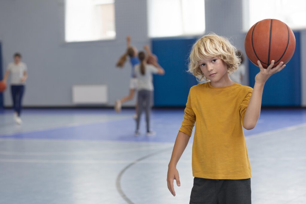 Ein Junge mit blonden Haaren und gelbem T-Shirt hält einen Basketball und schaut genau in die Kamera, im Hintergrund weitere Kinder die Basketball spielen