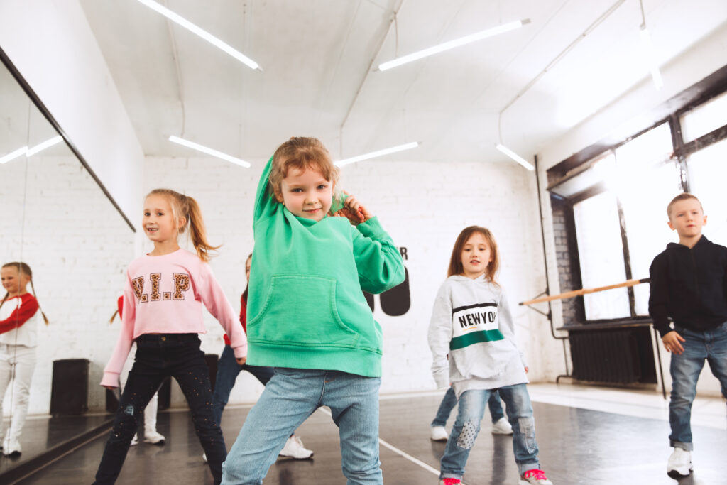 Vier Kinder, davon 1 Junge und 3 Mädchen, bewegen sich synchron zu Musik, im Hintergrund sind weitere Kinder