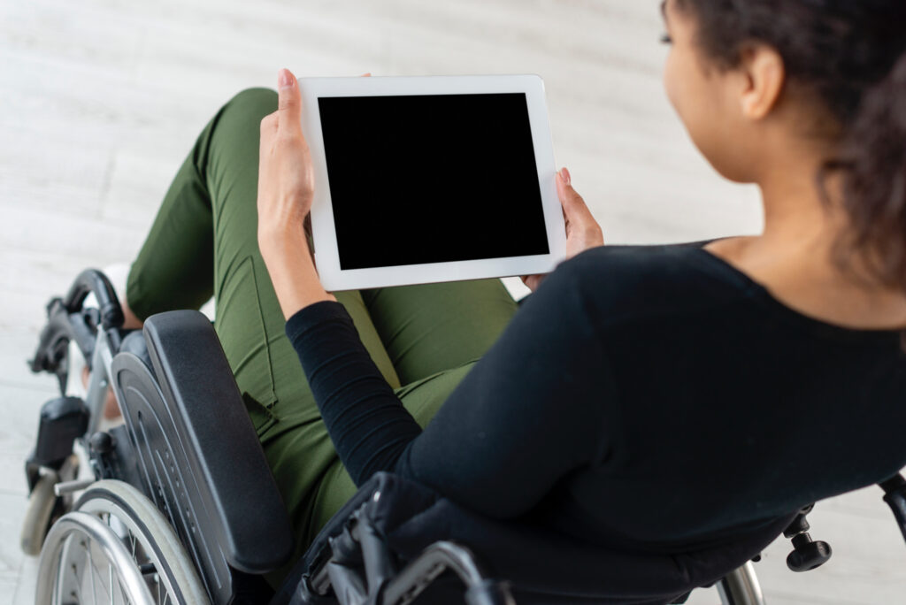 Eine junge Frau mit braunen Haaren im Rollstuhl hält ein Tablet in ihren Händen, das Bild wird über ihrer Schulter aufgenommen