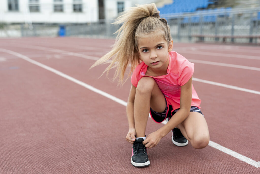 Ein junges Mädchen mit blonden Haaren kniet auf einer Kunststoffbahn in einem Sportstadion und bindet sich die Schuhe