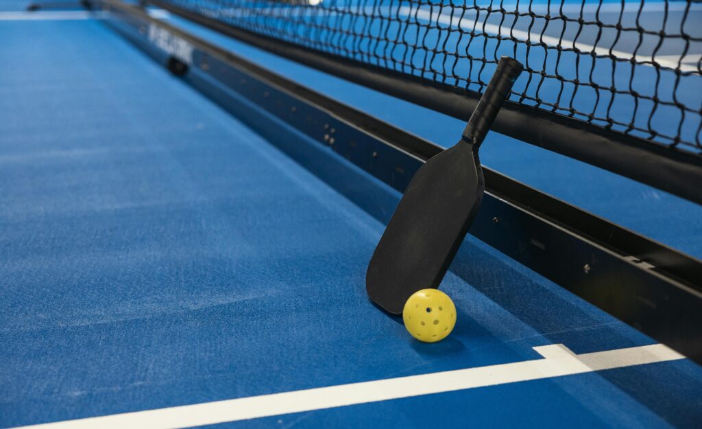 Ein blaues Spielfeld mit einem schwarzen Pickleball-Schläger und einem gelbem Pickleball-Ball in einer Nahaufnahme